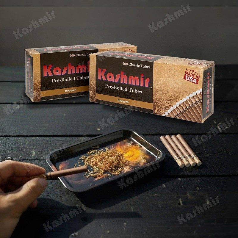 Two Kashmir Bronze pre-rolled tubes 200-count cartons on a dark table; a hand fills a brown tube over a tray of loose tobacco with four tubes set beside it.