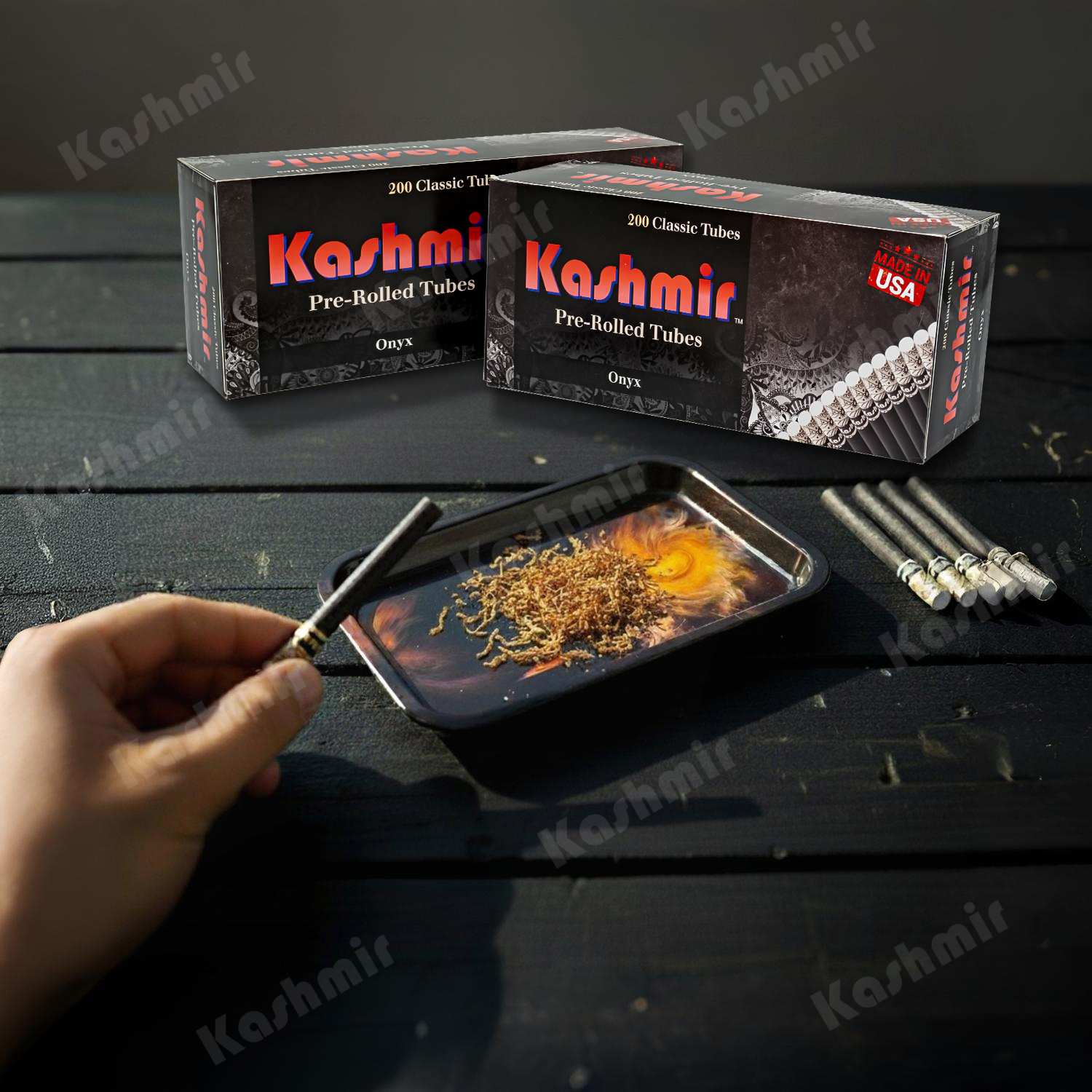 Two cartons of Kashmir Onyx rolling paper tubes, 200-count each, on a dark table while a hand fills a black tube over a small tray of loose legal hearbs; spare black tubes at right, Made in USA badge visible.