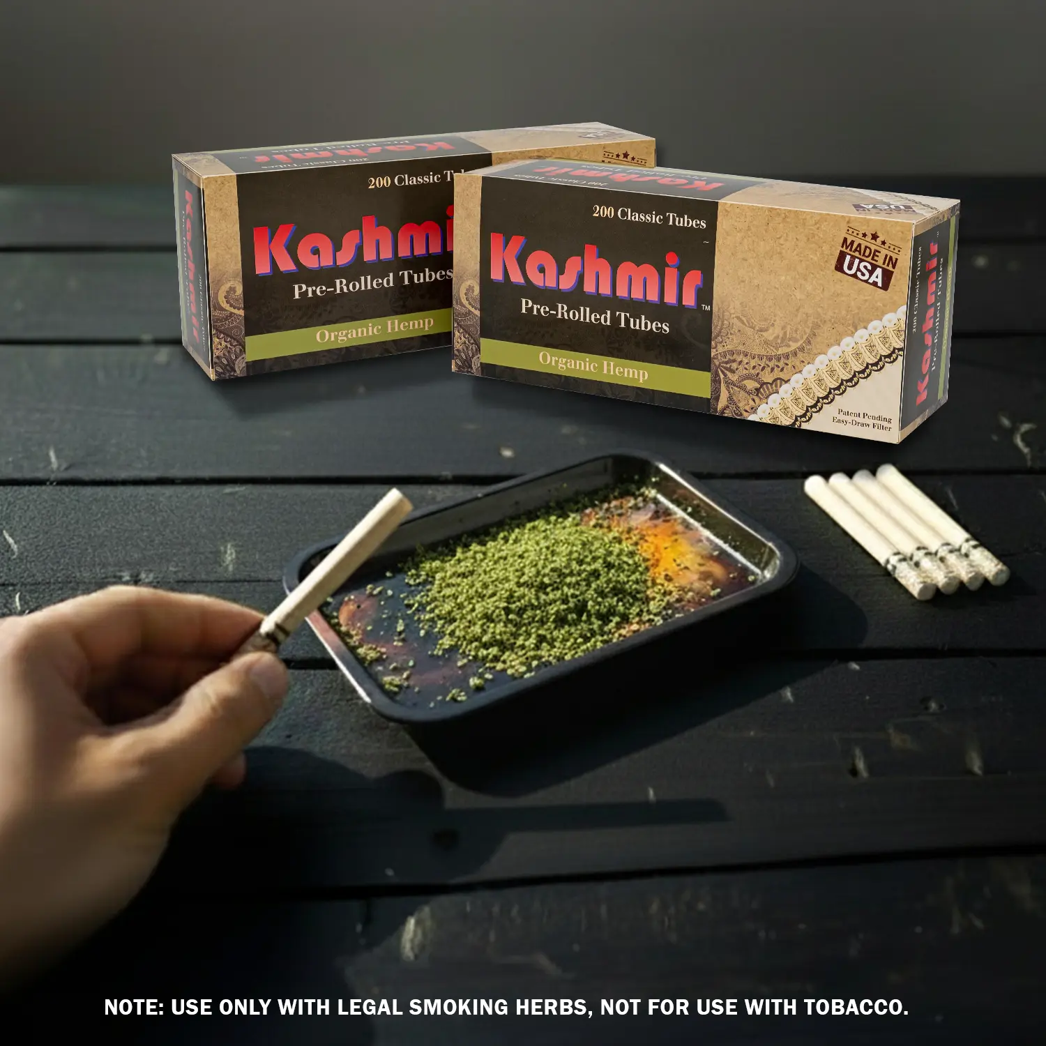 Kashmir Hemp Tubes — lifestyle scene with hand holding Organic Hemp Pre Rolled Filtered Tubes, rolling tray filled with leagal smoking herbs, two box of 200ct cartons and lined loose empty cigarette tubes (feature graphic).