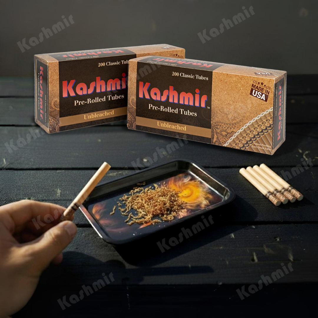 Two cartons of Kashmir Unbleached pre-rolled cigarette tubes, 200-count each, as a hand fills a natural filter tube over a tray of loose tobacco.
