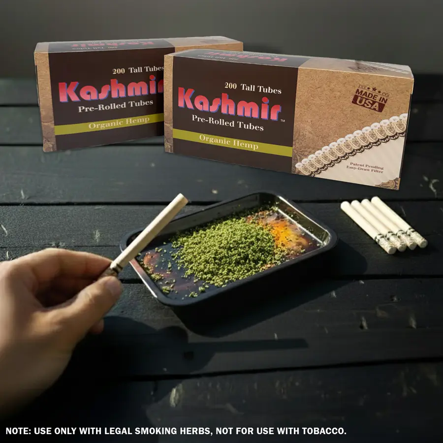 Two cartons of Kashmir Organic Hemp pre-rolled tubes (200-count each) on a table; a hand fills a hemp tube over a tray of loose herbs, with spare tubes nearby; Made in USA badge visible.